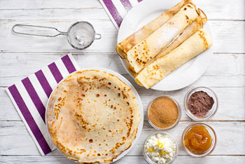 flat and rolled up pancakes on separate round plates, jam, cocoa powder, cinnamon in small glass bowls