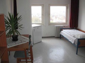 a basic hotel room interior with a single bed, a fridge, a table and a chair