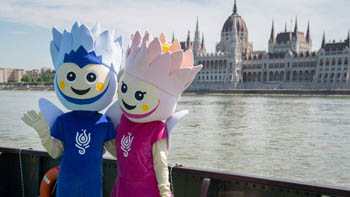 2 figure dressed in the mascot costume of the FINA: a blue and a pink water lily, the Parliament on the other side of the Danube in the background