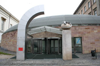 the grey domed entrance of the museum with 2 revolving doors, a a stone column with a gecko statue hugging a globe on top 