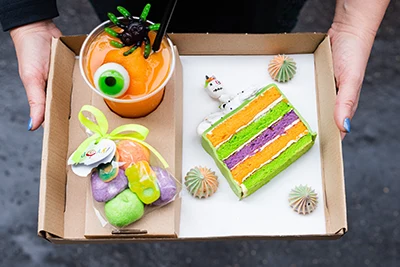 3 Halloween-themed sweets on a card board tray, a slice of cake, colorful candies in a cellophane bag, and a pumkin smoothie with a green eyeball and a black and green spider on top