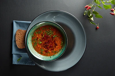 Bean goulash in a green soup bowl, a slice of whole wheat bread placed on the left side on a blue napkin