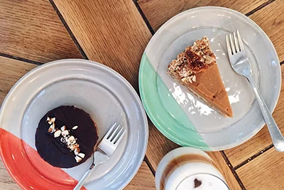 A slice of round chocolate covered cake on red /white plate, and a slice of cake with caramell or walnut icing on a green/white plate placed next to each other on a wooden table