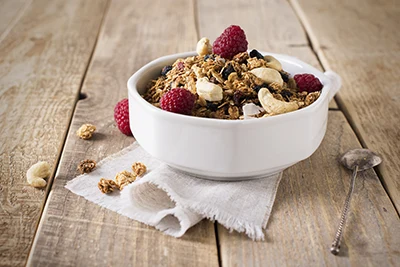 granola in a round, white ceramic bowl, with a few pieces of fresh raspberry on top, it's placed on a wooden table made of natural wood planks