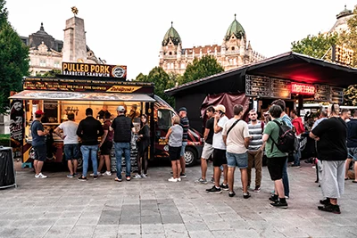 festival visitors standing in line at a wooden booth selling pulled pork street food at Liberty Sqr. The Russian Monument, and a twin-domed grand building can be seen in the background