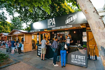 3 people at the wooden booth of IPA Beer House beer maker, under the canopy of platan trees on the Beer  Festival in Budapest