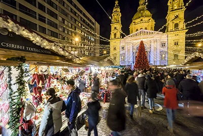 Christmas Market by Budapest Basilica: front view of the church illuminated at night with a red-lighted Christmas tree on the square and people strolling among the wooden booths