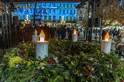 A large Advent Wreath with 3 candles lighted in Vörösmarty Square, the facade of Gerbeaud Cafe with festive light show on it in the background