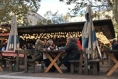 groups of peple eating at wooden tables covered with red and white checkered table cloth, strings of fairy light in the background