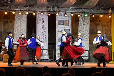 3 couples dancing in folk costume on a stage, men are wearing: white shirt, blue vest, black pants, women: embroidered red blouse and skirt, black handkerchief