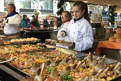 Roasted and grilled dishes in large serving trays and a young lady in white chef coat serving food for a visitor