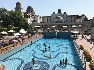 The outdoor wave pool of the Gellért Bath in summer with a handful of bathers in it. The domed buildings of the bath and hotel in the background.