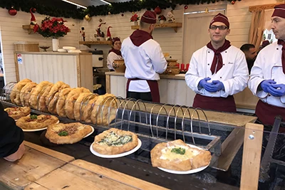 around a dozen langos cooling on a wire cooler, and some with toppings on plates, 3 male serves in white chef coat and dark red cap standing behind the counter