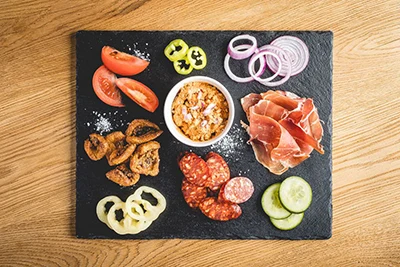assorted Hungarian cold cuts ,pork cracklings, cottage cheese spread in a white bowls, tomato slices, red onion, cucumber slices, paprika slices arranged on a black granite plate