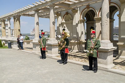 a trumpeter in hussar costume performing in front a colonnaded arcade up in Buda Castle, 2 more hussar musicians stand next to him on the left and right side