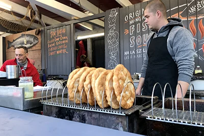 around half a dozen lángos on a stainless steel dripping tray, a young men withcropped dark hair in black apron stands behind the counter at the fair at the Basilica