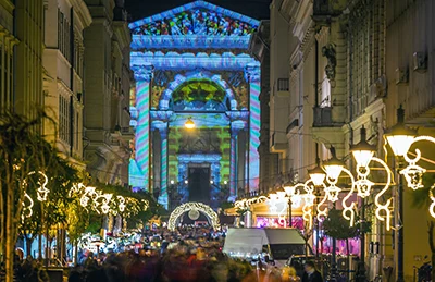 Christmas-themed light painting on the facade of Budapest's Basilica, lots of people at thefestive market on St. Stephen's Square in the evening, we can see the lamps in the street leading up to the church lit up