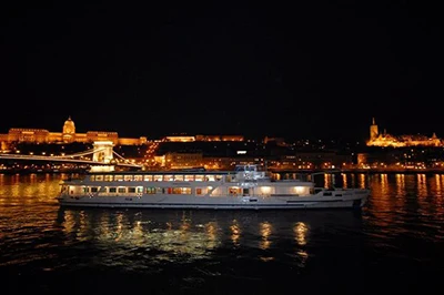a white cruise ship on the danube at night, Buda Castle and the Chain bridge illuminated in the background