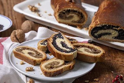 5 slices of beigli with poppy seed and walnut filling placed on a round white plate, two beigli rolls on a square white plate - one with walnut the other with poppy seed filling - in the background