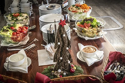 A festive wooden buffet table with cold salads, dressings, white plates and cutlery, a small wooden Christmas tree tbale ornament in the forefront 