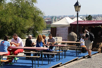 people sitting on wooden benches of Buda castle's terrace, eating, and sipping wine, the view of Pest in the background