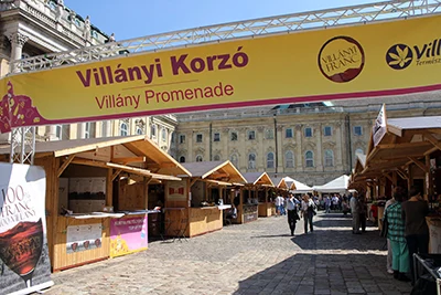Villány Promenade written in dark red on a large yellow ribbon, wooden booths in the courtyard