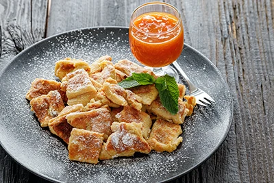 Kaisershmarnn sprinkled with icing sugar on a grey round plate placed on a dark brown rustic wooden tabletop, apricot jam in a small glass placed on the plate as well