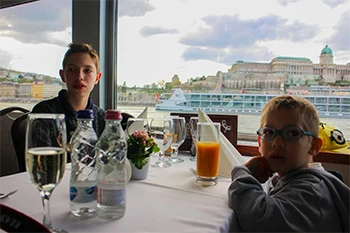 Our 11-yr and 6-yr old boys sitting at a table in a panorama cruise ship. Buda Castle can be seen through the window in the background on budapest cruise with lunch