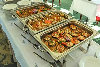 3 square shape stainless steel food containers filled with stuffed cabbage, beef stew and grilled aubergine in marinara sauceon a white clothed buffet table