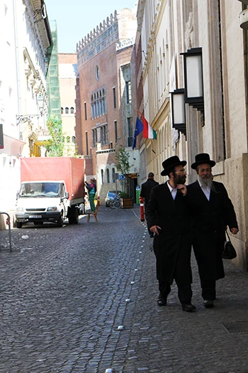 A photo of two middle-aged orthodox Jewish men dressed in traditional balck suit, white shirt and wearing a black hat walking in kazinczy Street in Budapest. The red-bricked synagouge of the same name as the street can be seen in the background.