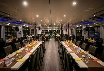 The interior of the Ctamaran cruise boat where the Budapest fireworks cruise will be hosted. Two rows of elegantly set dinner tables (chocolate brown running table cloth, yellow and orange napkins, silverware, wine glasses, flower arrangements) with ambient blue and white lighting.