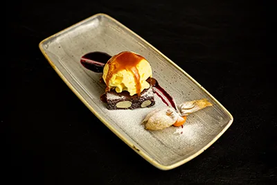 a square slice of dark choc brownie with cashew, with a scoop of vanilla ice cream on top, served on a long square shaped beige plate