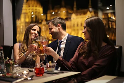 2 young women and a men clinking wine glasses on a late night Danube cruise, the blurred view of the Hungarian Parliament in the window