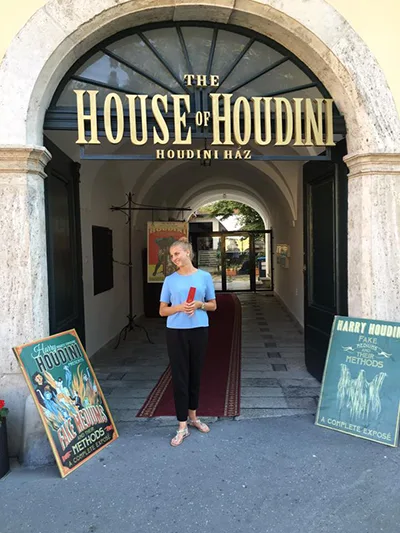 A young female hostess in black pants and blue t-shirt standing at the vaulted entrance of teh House of Houdini