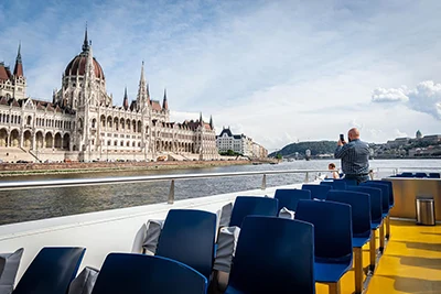 A man taking photos of the Parliament building, from the open deck of a cruise boat during the day.