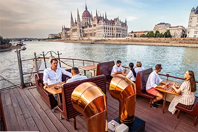 Passengers sitting on the upper opne deck of the Kisfaludy historic paddle steam boat. The Parliament in the background.