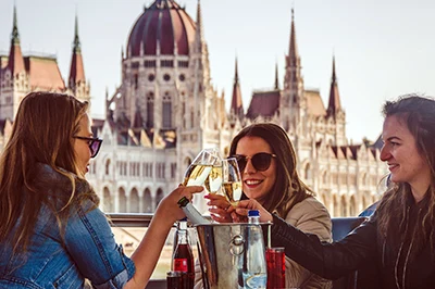 3 young women clinking glasses of prosecco on a cruise ship in Budapest during the day. The Parliament building can be seen in the background.