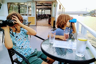 two young boys with binoculars sitting at a table on the open deck of the cruise boat on the Danube in Budapest