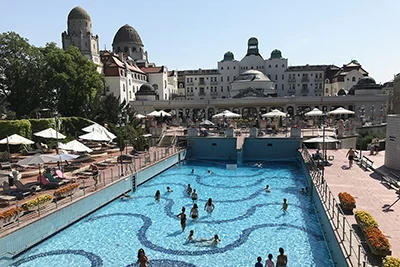 The outdoor wave pool of the Gellért Bath in summer with a handful of bathers in it. The domed buildings of the bath and hotel in the background.