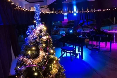 the dinner hall of a cruise ship with dimmed blue lighting, a small stage with a piano and a Xmas tree with the lights on in the foreground