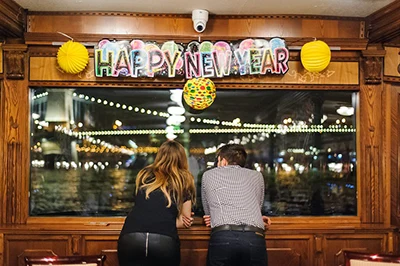 a young couple enjoying the night view of the Danube with the Chain Bridge all lit up through a panorama window of a cruise ship, "Happy New Year" decor on the top window frame
