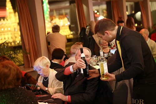 a waiter serving a bottle of wine for the guests sitting at their tables on a festive dinner cruise in Budapest