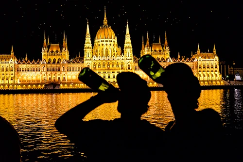 two men in baseball cap drinking prosecco from the bottle and looking at the Parliament building on a Danube cruise party
