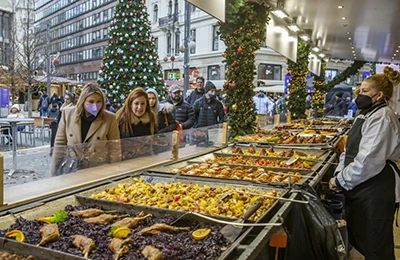 tourists looking at the food offer at one of the booths on the Christams Market in Vörösmarty tér, the Xmas tree can be seen in the background