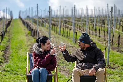 a young woman with black hair and aman wearing a cap and sunglasses clinking with a glass of wine sitting in a vineyard