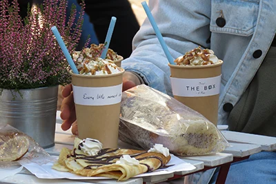 2 coffee drinks in abrown paper cup (fancy latte with whipped cream and syrup), a chimney cake and a pancake on apaper plate