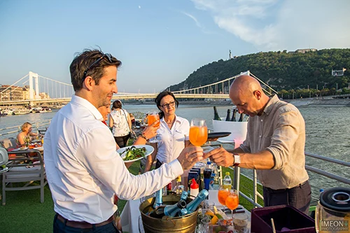 host handing a glass of orange colored drink to a middle-aged man on the open terrace of a boat, Elizabeth bridge and Gelléert Hill in the background in daytime