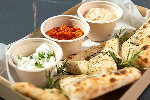 Foccacia bread pieces with 3 dips in paper dishes on a paper box tray