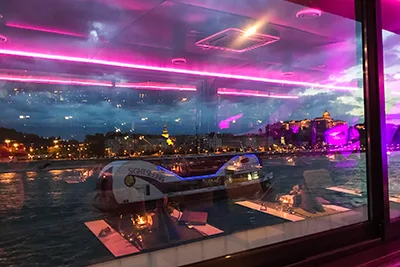 View of the Buda side through apanorama window of a river boat at dusk, the interior and pinkish-purple lighting of the baot is reflected in the window glass