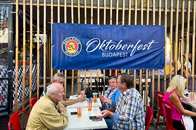 4 older men sitting at a table , a blue banner with Oktoberfest Budapest written hanging next to them on a wooden divider